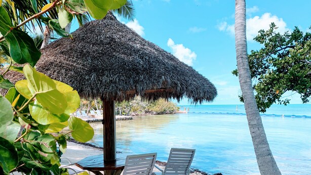 A beachside setting with a thatched umbrella, two lounge chairs, and a wooden table. - Private Island, FL Keys Entire Island, Home, A/C, Boat, Dock, Beach, Bikes, WiFi (Marathon)