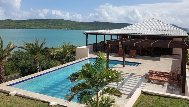 Une piscine avec vue sur la mer, un espace salon couvert et un bâtiment doté d’un toit en tuiles.