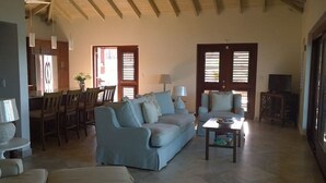 Un vaste salon avec un canapé bleu clair, un fauteuil assorti et une table basse. On y trouve également un coin repas avec des chaises et une table en bois, ainsi qu’une cuisine à l’arrière-plan.