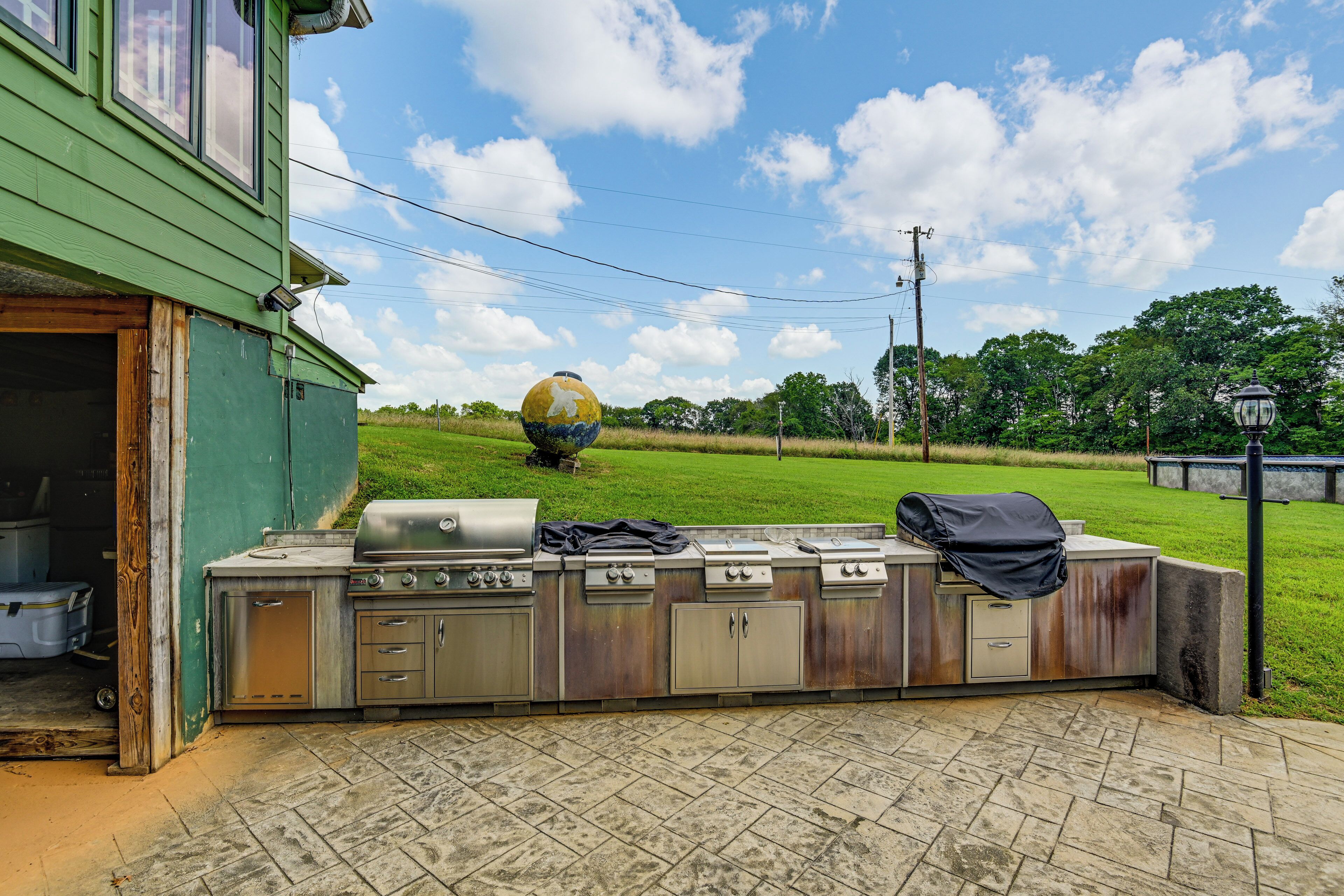 An outdoor kitchen with a grill and storage cabinets, a green building in the background, and a grassy field with a large spherical sculpture.