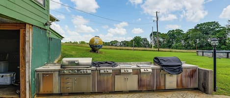 An outdoor kitchen with a grill and storage cabinets, a green building in the background, and a grassy field with a large spherical sculpture.