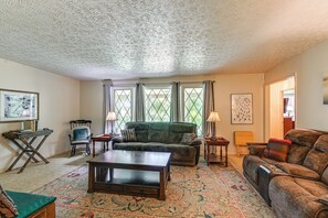 Uma sala de estar com um sofá verde, uma mesa de centro de madeira e um tapete estampado. Há duas janelas com cortinas, uma cadeira e uma pequena mesa com abajur.