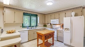 A kitchen with white cabinets, a wooden table, a refrigerator, a microwave, and a window with green curtains.