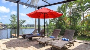 Un espace extérieur aménagé, abrité et doté de fauteuils de jardin en osier et d’un parasol rouge, offrant une vue sur une étendue d’eau et des palmiers.