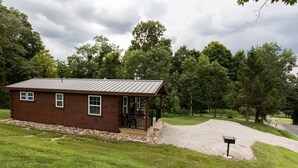 A small, single-story wooden cabin with a metal roof, a porch with a railing, and a gravel driveway.