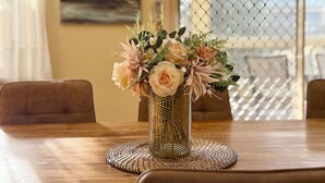 Een eettafel met een geweven placemat, een vaas met bloemen en uitzicht door een hekwerk.