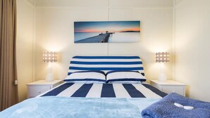 Hotelkamer met een bed met gestreept beddengoed, twee bedlampen, een schilderij van een ondergaande zon boven water en uitzicht op een pier.
