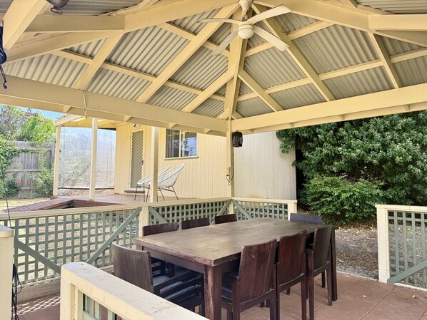 A covered patio with a wooden table and chairs, a ceiling fan, and a view of a garden.
