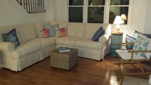Uma sala de estar com um sofá seccional bege, uma mesa de centro de vime, uma cadeira de madeira e um abajur.