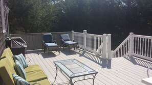 Relax on the covered deck with lounge chairs, a table, and barbecue grill, surrounded by trees and a building facade.
