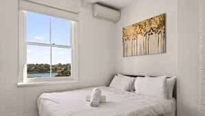A bedroom with a large window, a bed with white linens, a painting on the wall, and an air conditioning unit. - Waterview terrace in rock (Millers Point)