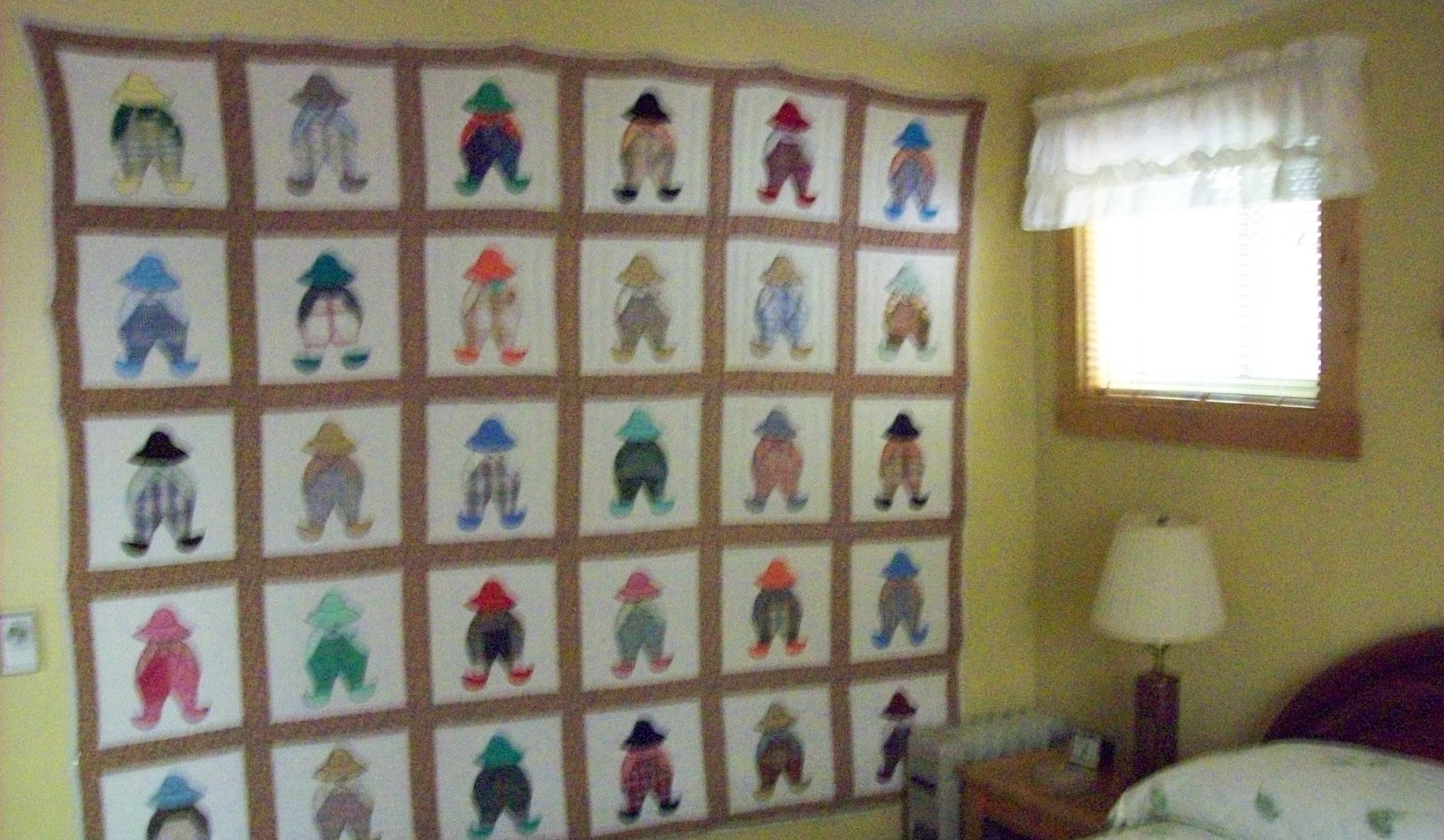 A room with a wall of framed artwork depicting various figures. There is a window with a white valance, a bedside table with a lamp, and a bed with a floral-patterned bedspread.