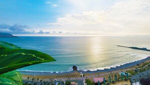 Vista costera con playa, océano y una carretera con coches aparcados.