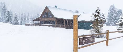 Un cartel de madera con la leyenda 'Birch Valley', una cerca de madera y un edificio cubierto de nieve al fondo.