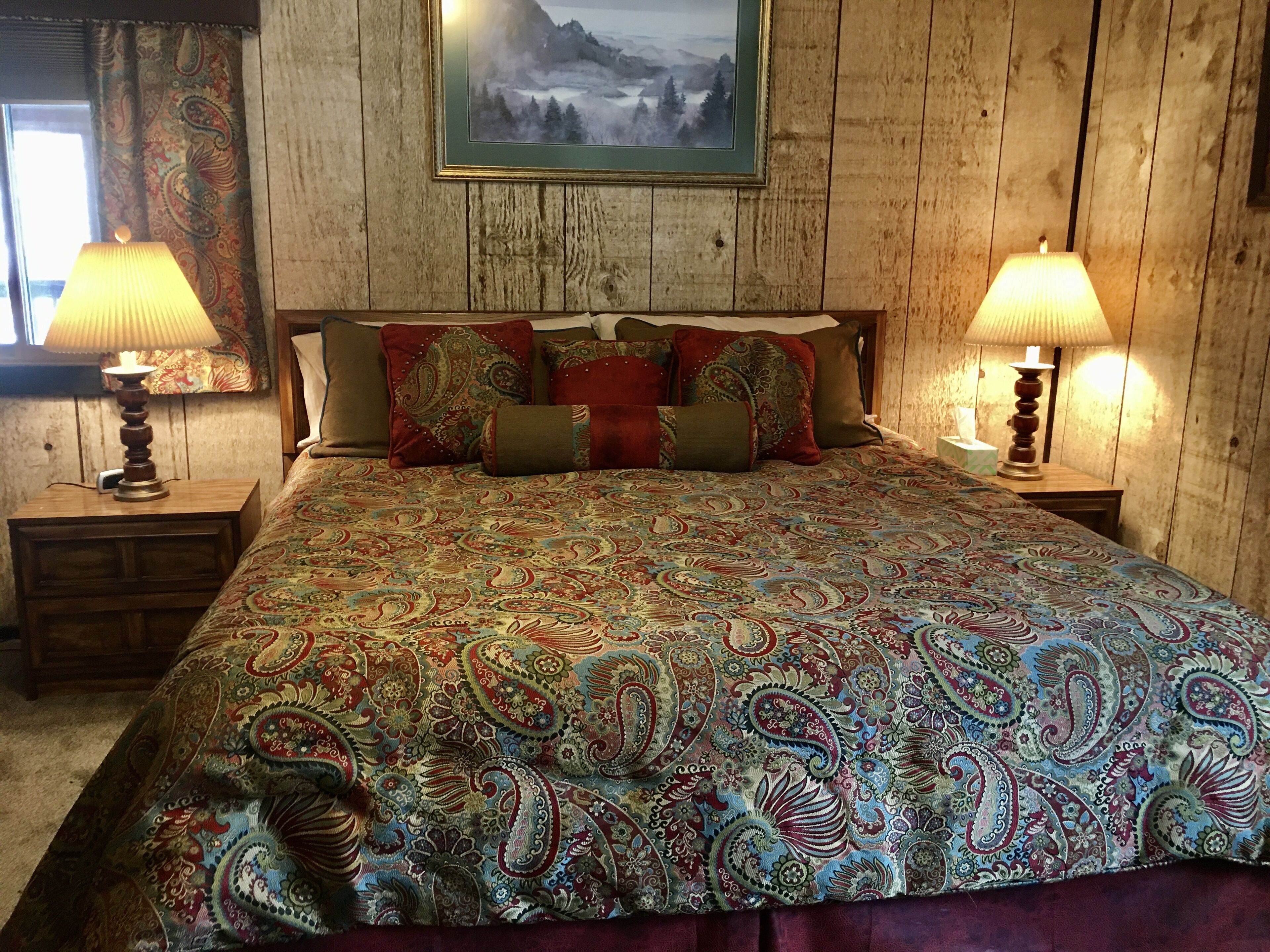 Une chambre à coucher avec un grand lit, deux lampes de chevet, une tête de lit en bois et un tableau représentant un paysage, encadré, accroché au mur.