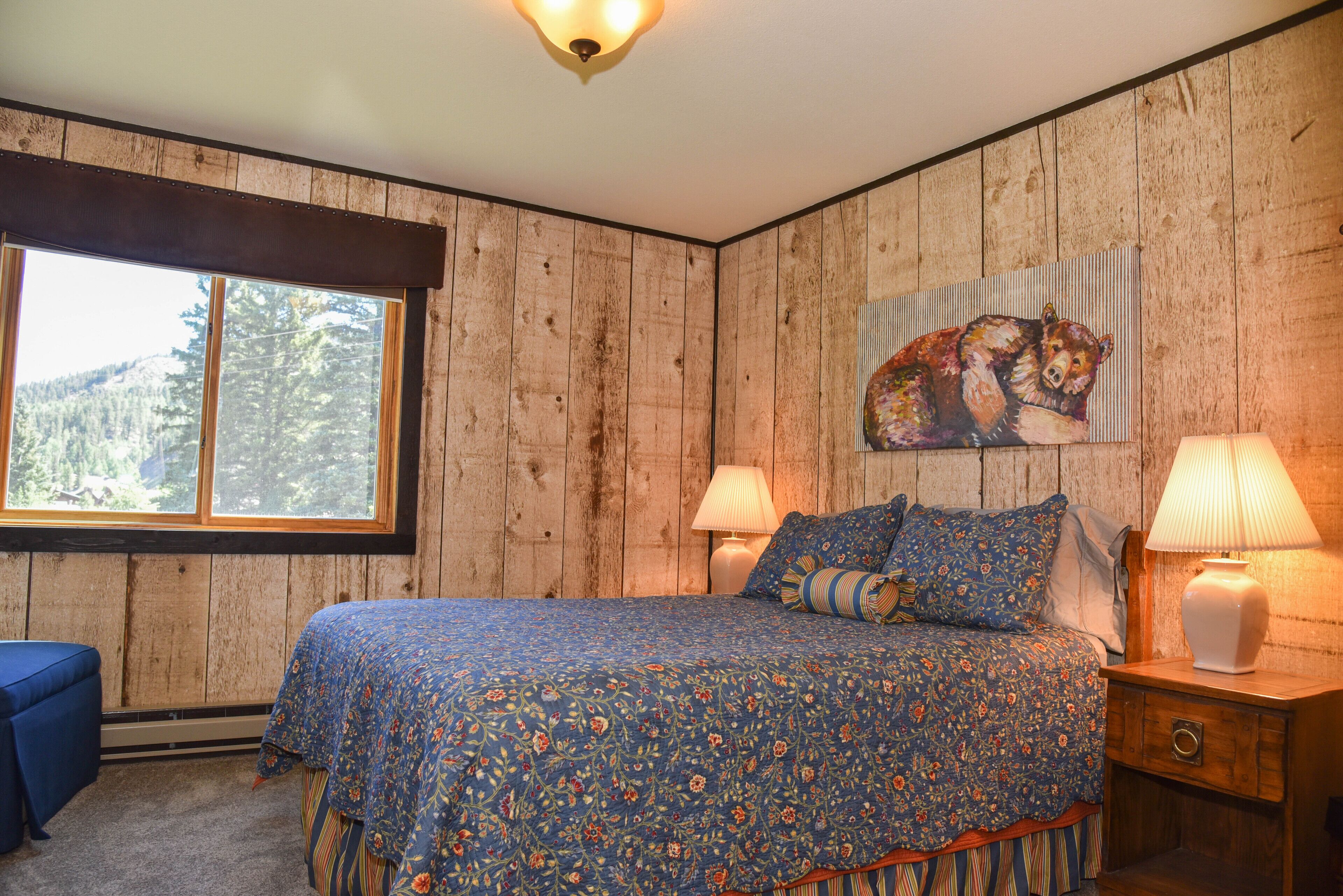 Een slaapkamer met een houten wand, een groot bed met bloemenbeddels, twee bedlampjes en een raam met uitzicht op bomen.