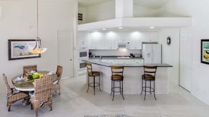 Een moderne keuken met witte kasten, een centraal kookeiland met barstoelen en een eethoek met rieten stoelen.