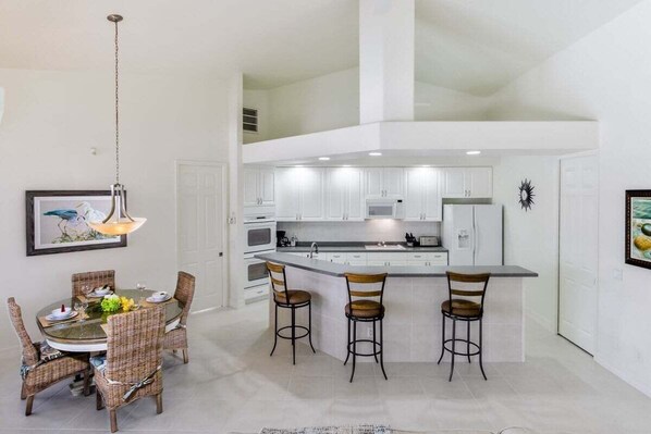 Une cuisine moderne avec des meubles blancs, un îlot central doté de tabourets de bar et un coin repas avec des chaises en osier.