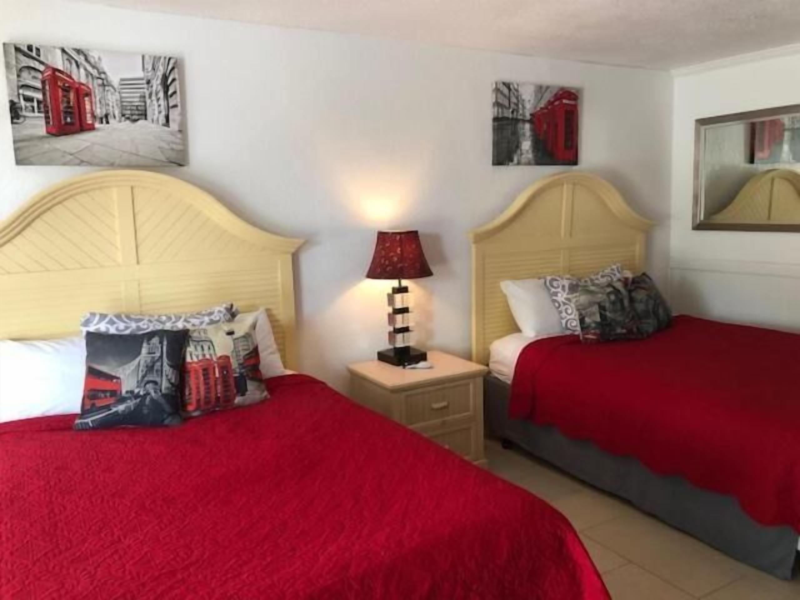 A hotel room with two beds, each with a red blanket and a pillow featuring a red telephone booth design. There is a nightstand with a lamp between the beds.
