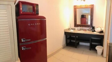 Un baño pequeño con un refrigerador Galanz rojo, un lavamanos con espejo y un cubo de basura.
