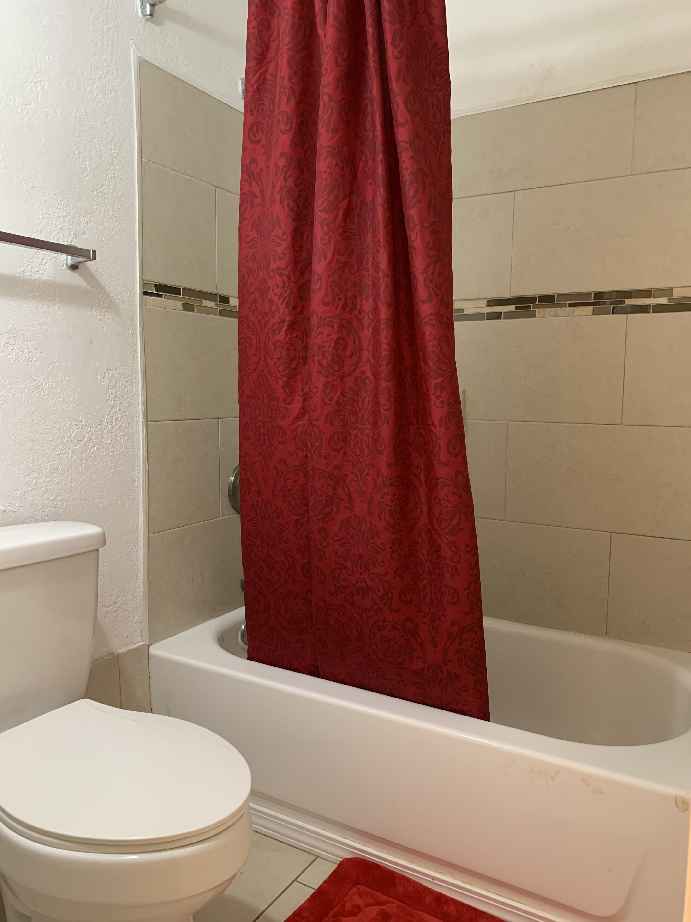 A bathroom with a white toilet, a bathtub with a red shower curtain, and beige tiled walls.