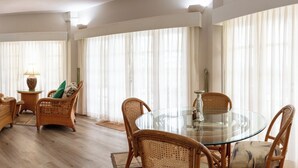 A spacious living area with a glass-top dining table surrounded by wicker chairs, a television mounted on the wall, and sheer curtains allowing natural light.