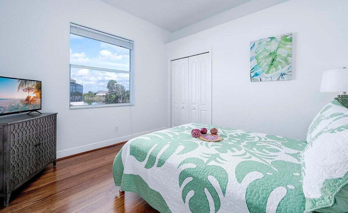 A bedroom with a large window, a bed with a green and white patterned bedspread, a TV on a wooden cabinet, and a white door.