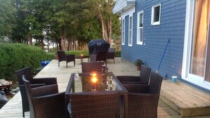 Eine Terrasse im Freien mit Korbmöbeln und einem Glastisch, umgeben von einem blauen Haus und Bäumen.