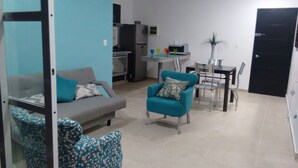 Un salon avec un canapé gris, un fauteuil turquoise et un pouf à motifs. Une table à manger avec des chaises se trouve à l’arrière-plan.