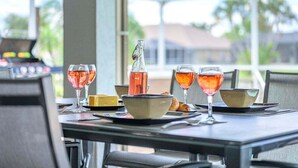 Ein Tisch ist gedeckt mit Weingläsern, einer Flasche und Schalen, mit Blick auf Palmen und Häuser durch eine Glasstruktur.