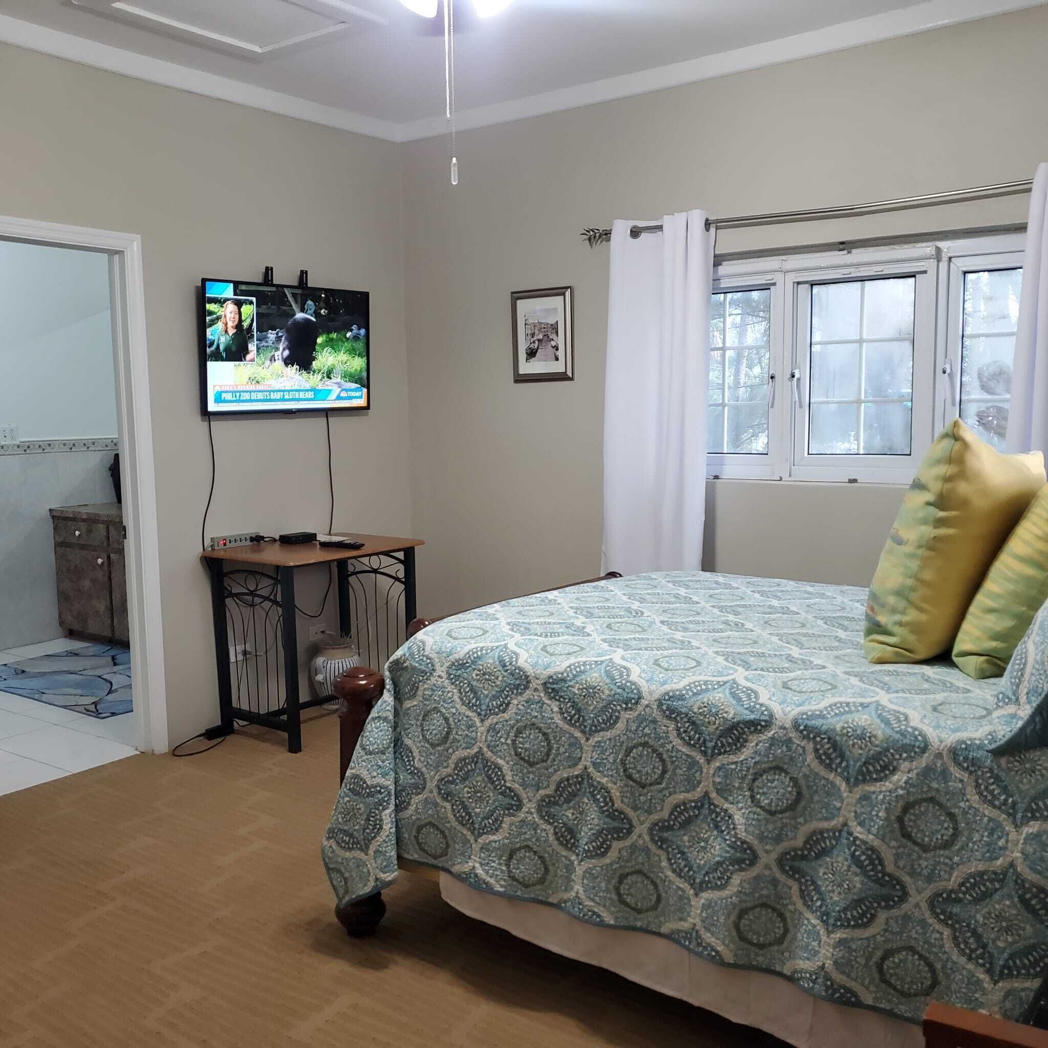 A bedroom with a bed, a TV mounted on the wall, a small table, and a window with curtains.
