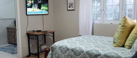 A bedroom with a bed, a TV mounted on the wall, a small table, and a window with curtains.