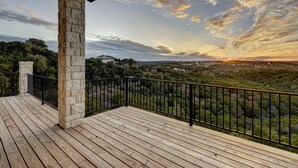 A wooden deck with stone pillars, a black railing, and a view of a landscape with trees and buildings.