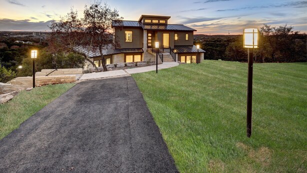 A modern two-story house with a paved driveway leading to the entrance, surrounded by a well-maintained lawn and trees.