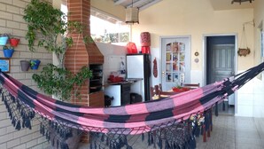 A spacious living area with a hammock, a brick fireplace, and a kitchen in the background. - Complete house in Praia do Rosa (Ibiraquera)