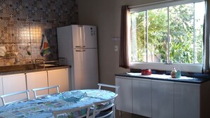 A kitchen with a tiled backsplash, a refrigerator, a dining table with chairs, and a window with a view of greenery. - Complete house in Praia do Rosa (Ibiraquera)