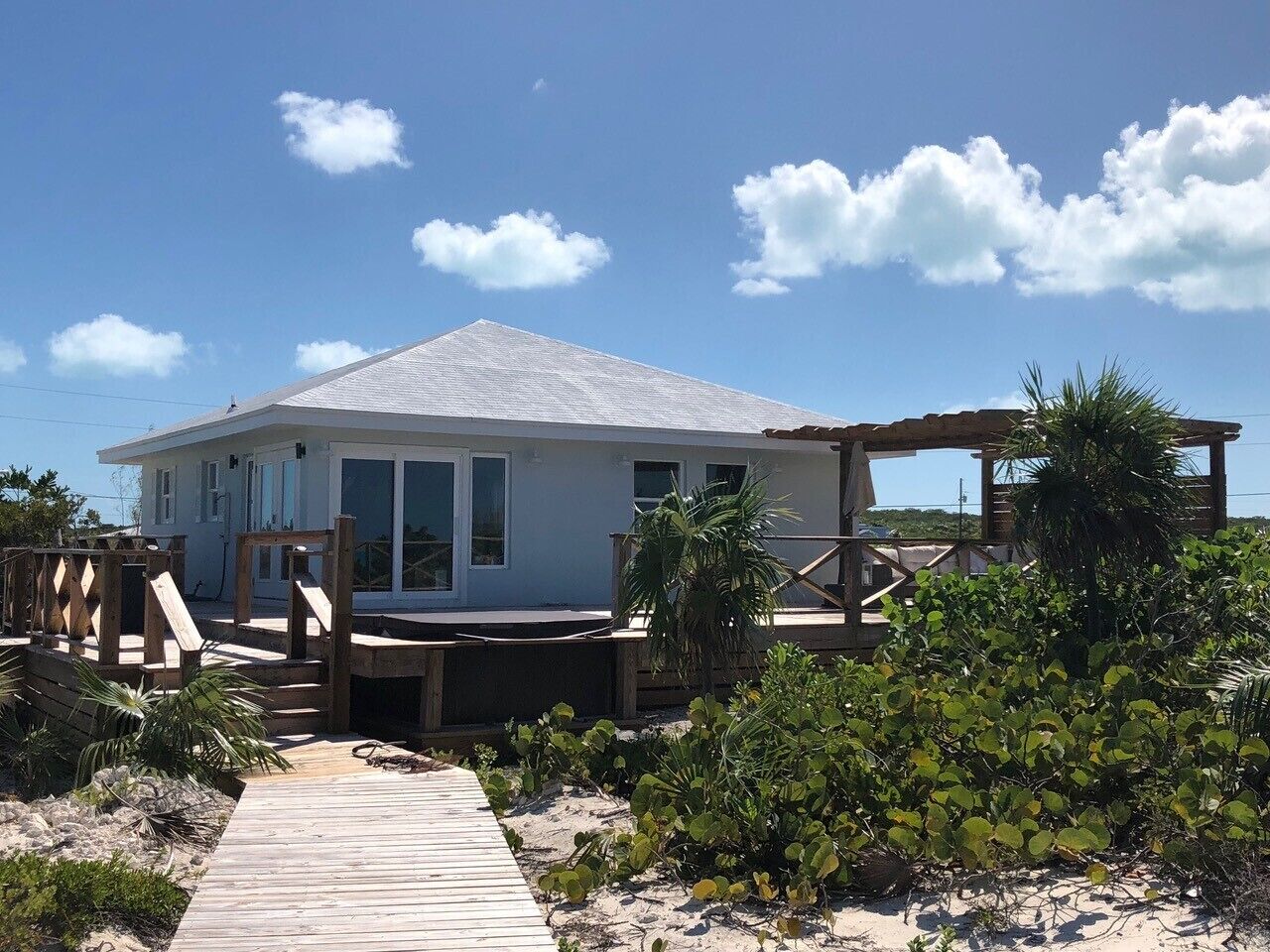 Ein Strandhaus mit Holzterrasse, überdachter Außenbereich und sandigem Weg zum Eingang.