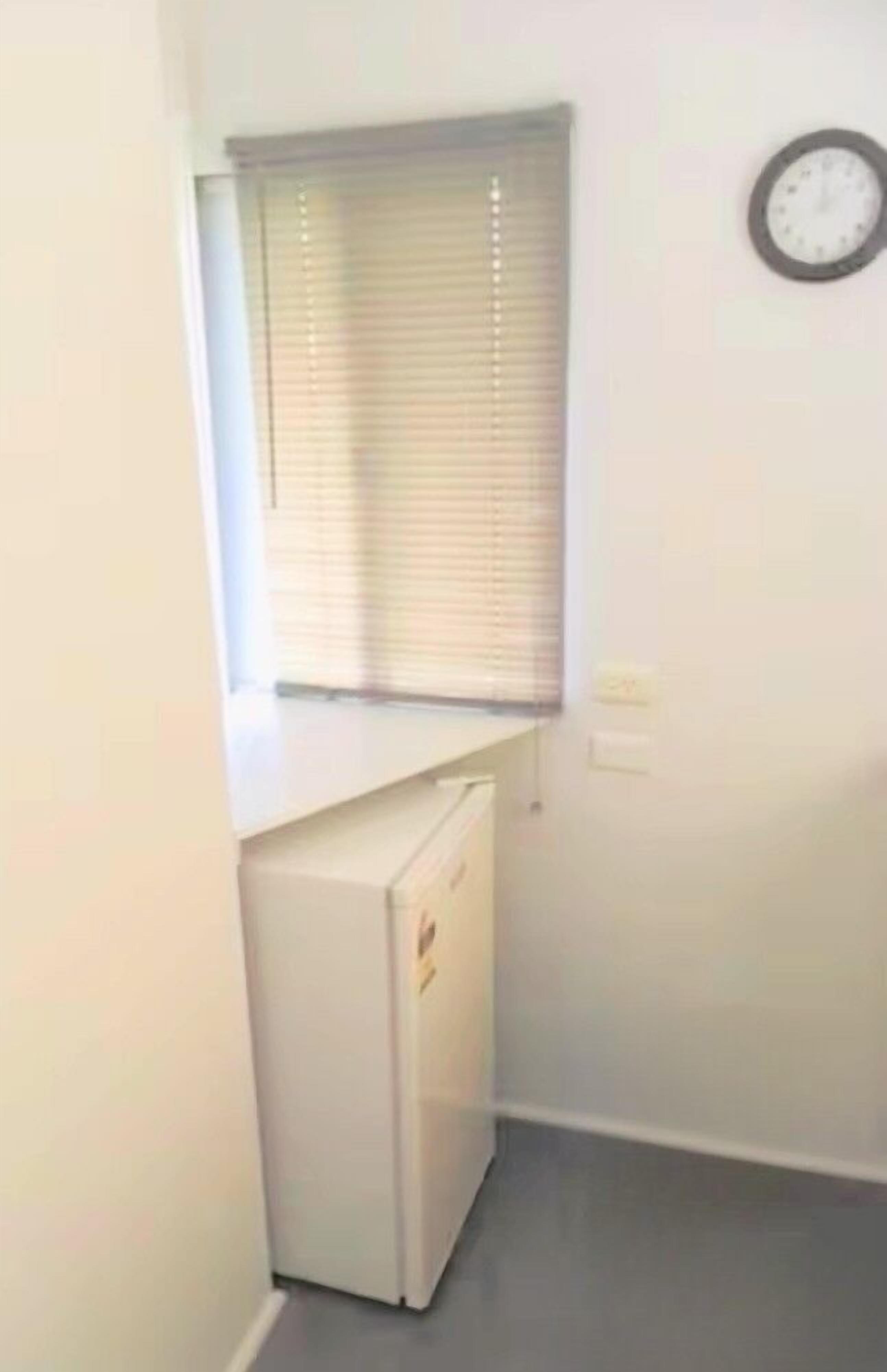 Una habitación pequeña con una ventana, un reloj en la pared y un electrodoméstico blanco.