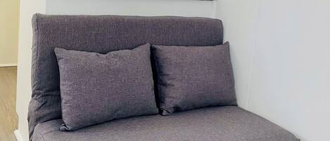 A gray sofa with wooden legs in a room with white walls, a yellow cabinet, and a window with blinds.
