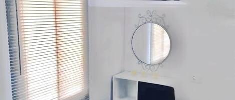 Un dormitorio con cama, televisor montado en la pared, ventana con persianas y mesita de noche con espejo y lámpara pequeña.