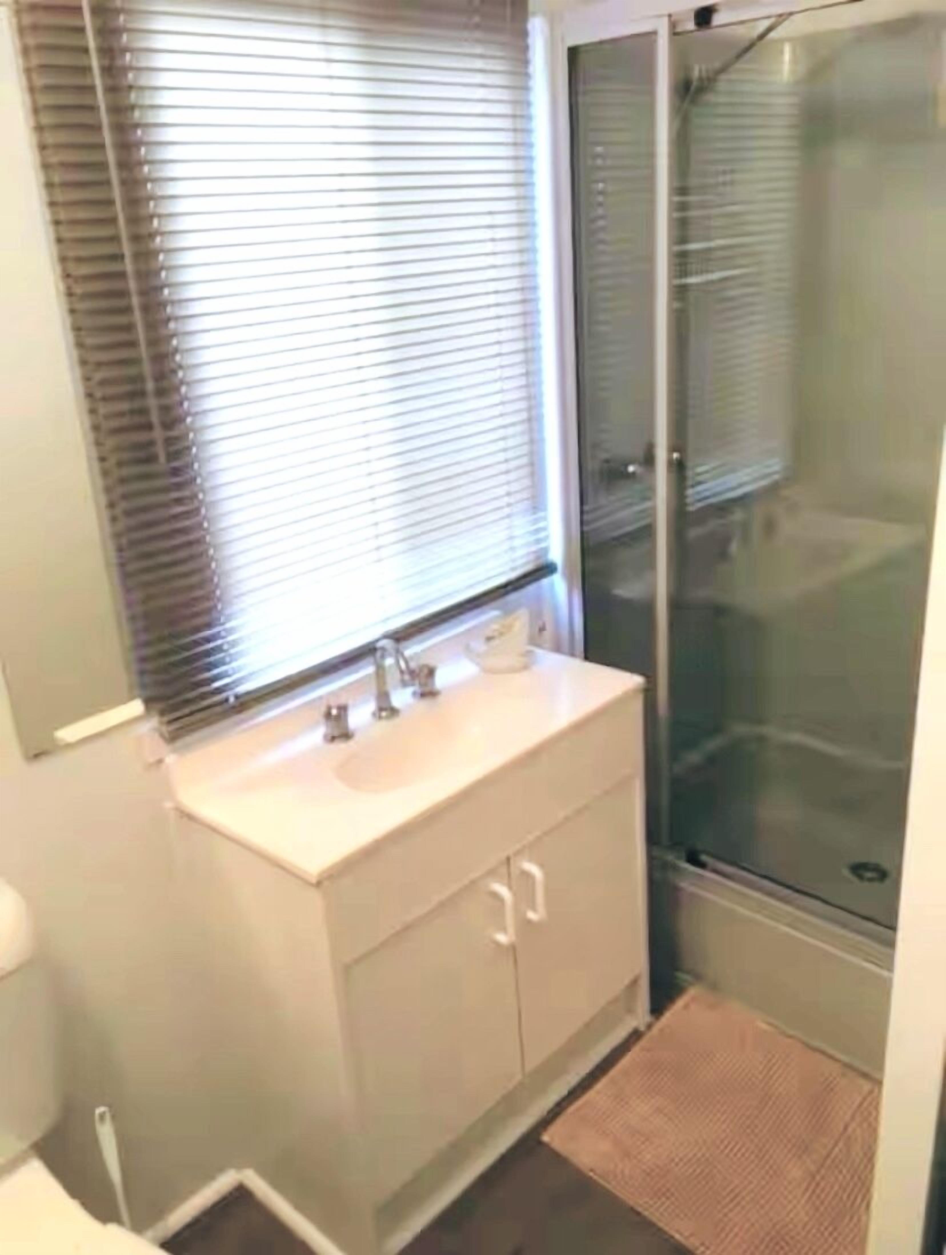 A bathroom with a glass shower enclosure, a sink with a mirror, and a window with blinds.