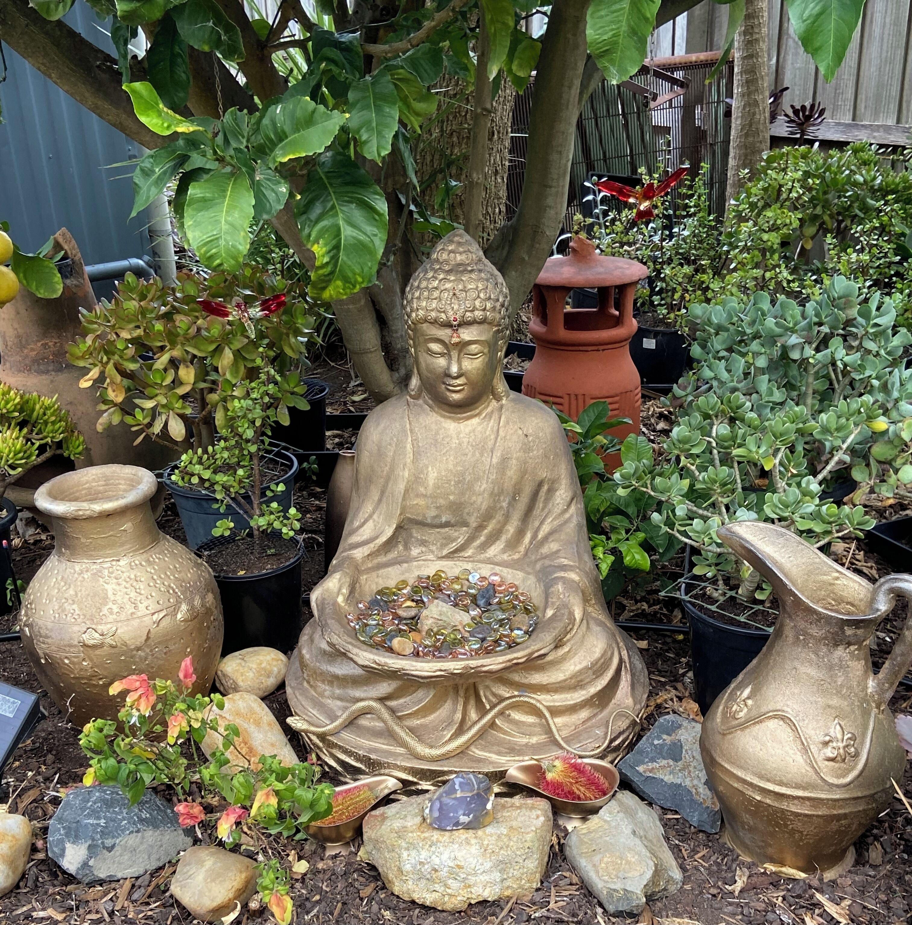A garden statue of a seated figure with a bowl in front, surrounded by potted plants and rocks.