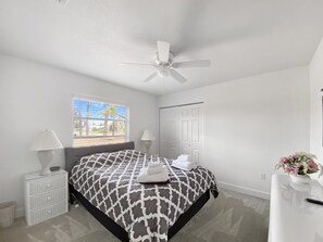 Une chambre Ă coucher comprenant un lit, un ventilateur de plafond, une fenĂŞtre avec des stores, une table de chevet, une commode et un vase avec des fleurs.
