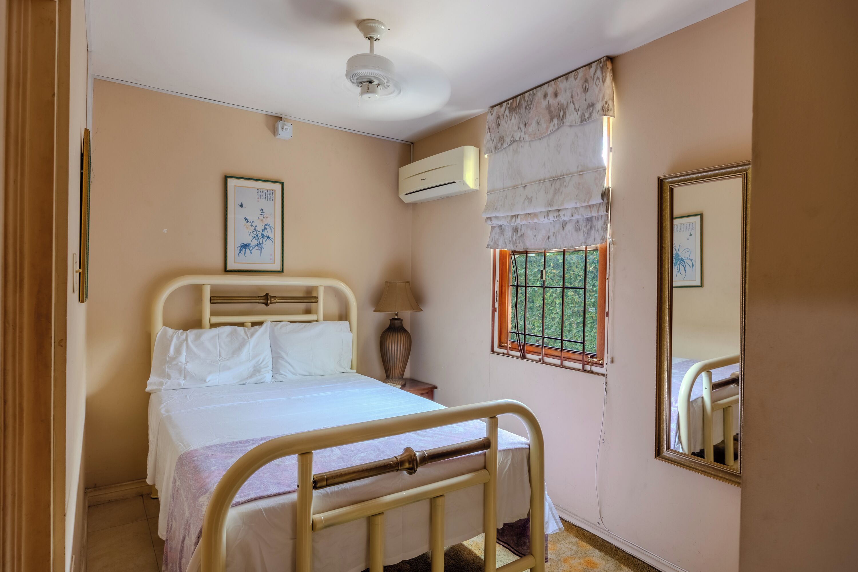 A bedroom with a bed, a bedside lamp, an air conditioning unit, a window with a view, and a mirror.
