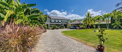 A paved pathway leading to a two-story house with a garage, surrounded by lush greenery and a clear blue sky.