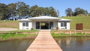 Een modern huis met een houten terras dat leidt naar een glazen voordeur, omringd door een gazon en bomen.