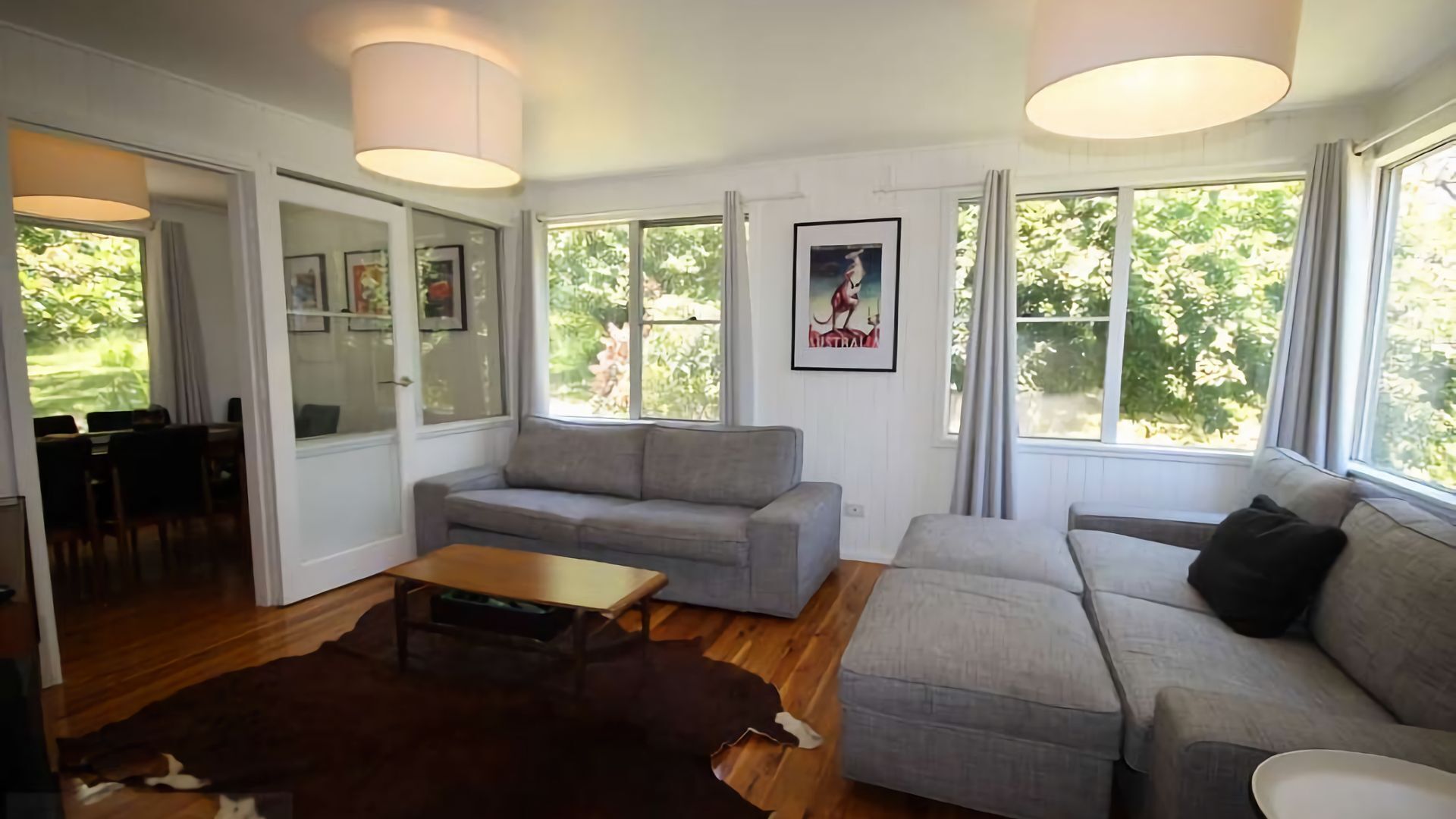 Ein geräumiges Wohnzimmer mit grauem Sofa, einem Holztisch und einem braunen Teppich. Große Fenster mit Vorhängen, eine Essecke mit Stühlen und gerahmte Bilder an der Wand.