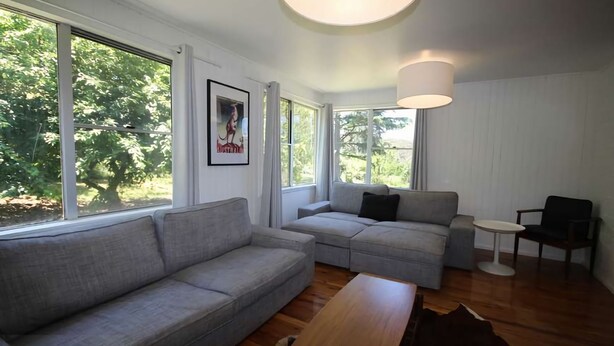 A modern living room with a grey sofa, a smaller grey couch, a wooden coffee table, and a black chair. There is a framed picture on the wall, a round table, and a large window with curtains. - Carawatha | Private location in Blackheath with a rural feel (BLACKHEATH)