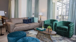 Een woonkamer met een beige bank, een groene fluweel fauteuil en een blauwe poef. Er staat een salontafel met een lamp en een kleine plant.