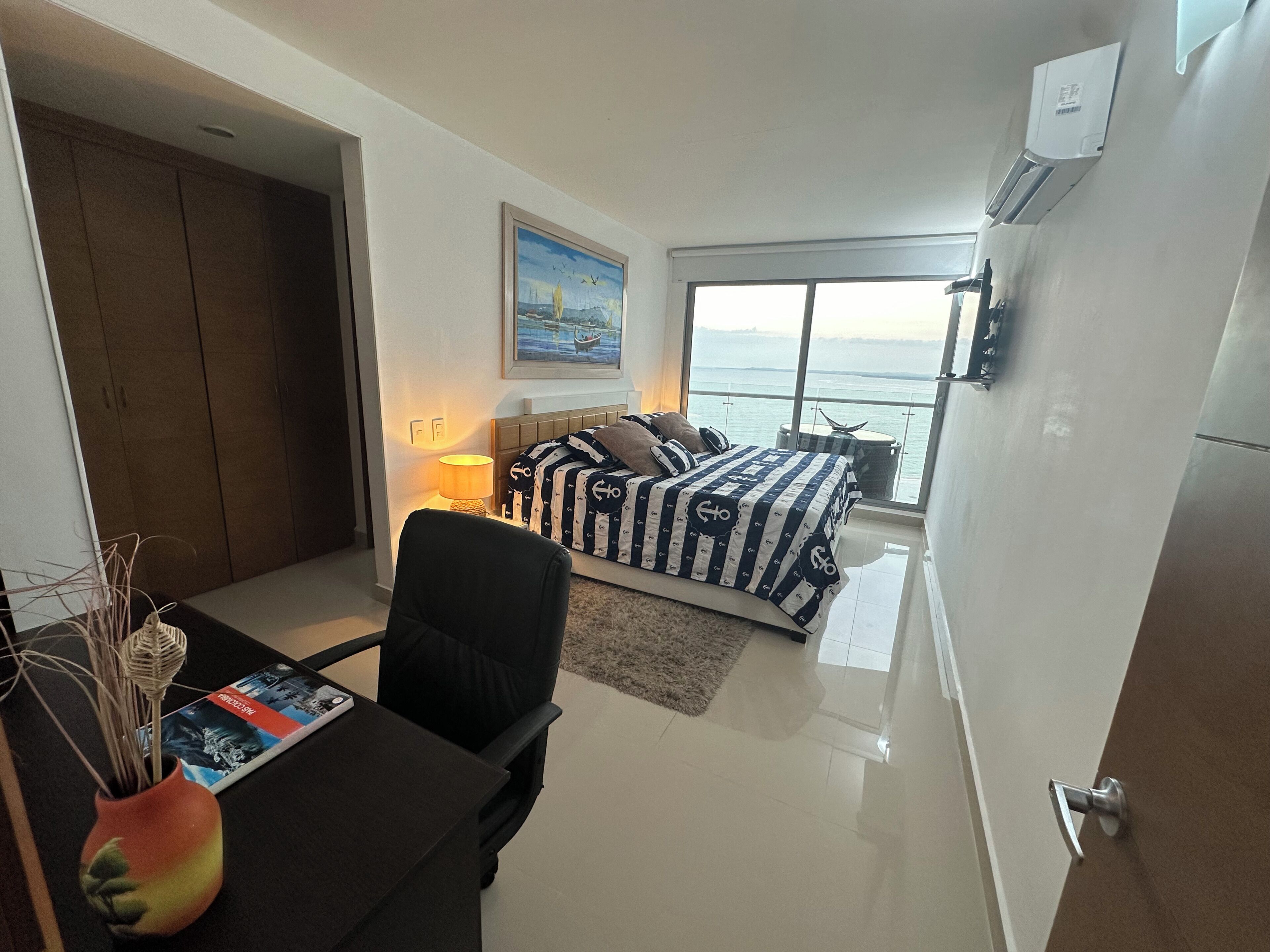 Une chambre à coucher avec un lit, un bureau et une chaise, un vase et une fenêtre donnant sur la mer.